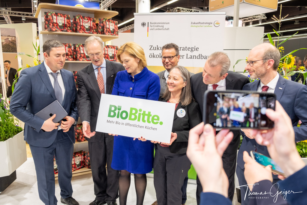 Julia Klöckner, Bundesministerin, hält ein Schild "BioBitte, Mehr Bio in öffentlichen Küchen", im Vordergrund ein Handy zum Fotografieren, mehrere Menschen darum herum widmen sich dem Schild